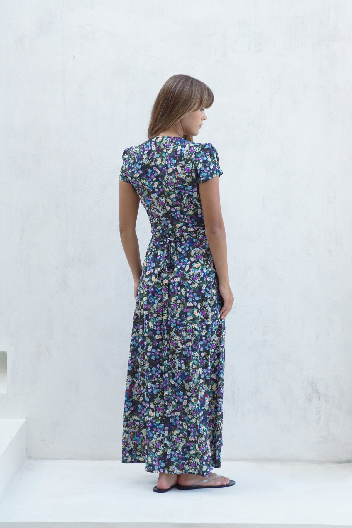 A woman wearing a short-sleeved maxi dress with a dense blue, purple, and green floral print, shown from the back against a white textured wall.