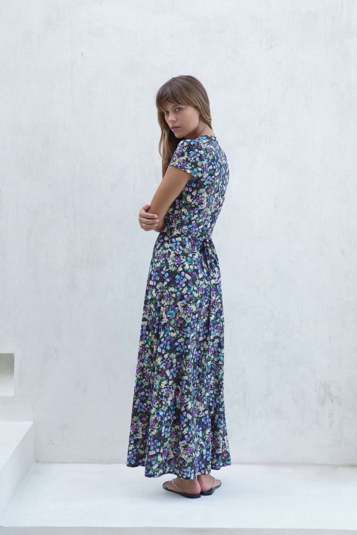 A young girl with long light brown hair stands on white steps against a white textured wall, wearing a short-sleeved maxi dress with a dense multicolor floral print on a dark navy background.