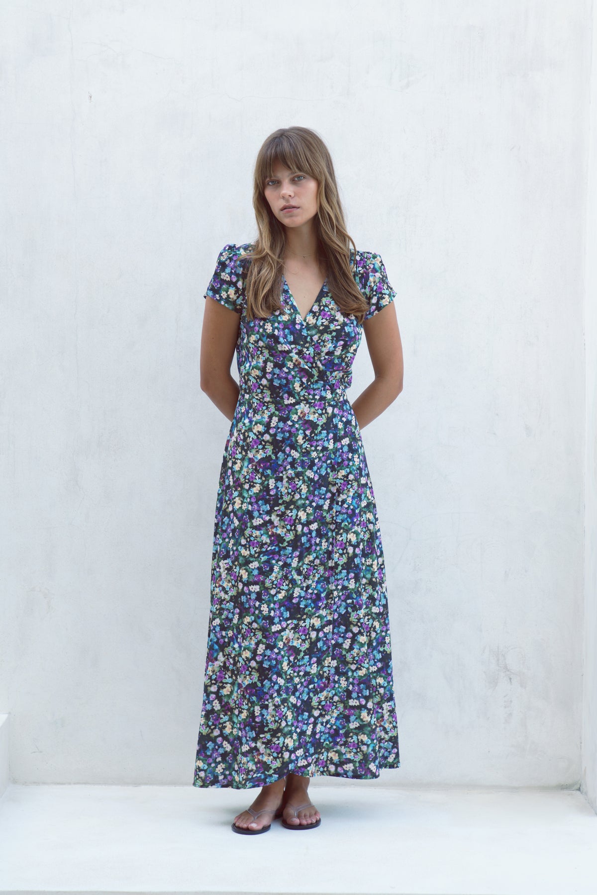 A woman modeling a short-sleeved, wrap-style maxi dress with a dense blue, purple, and green floral print, standing against a textured white wall.