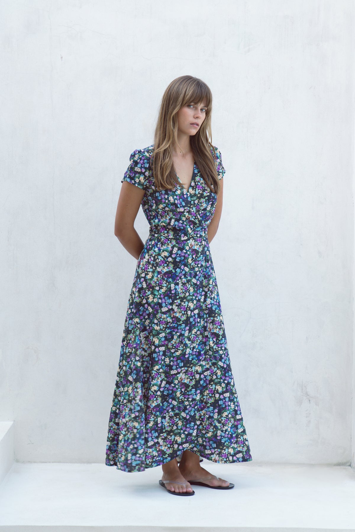 A woman modeling a short-sleeved maxi dress with a dense blue, purple, and green floral print, standing against a white textured wall.