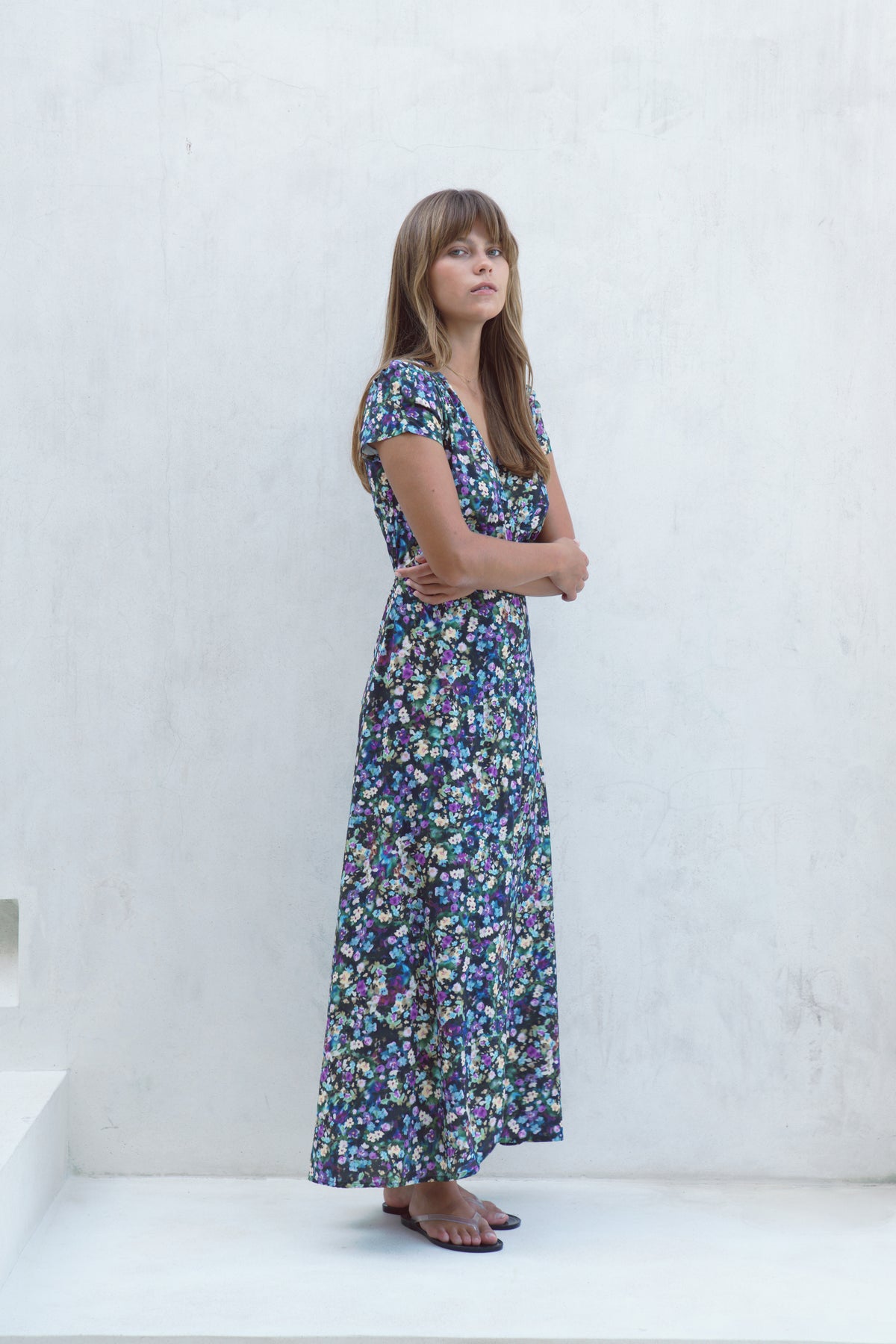 A woman wearing a short-sleeved maxi dress with a dense blue, green, and yellow floral print, standing against a textured white wall.