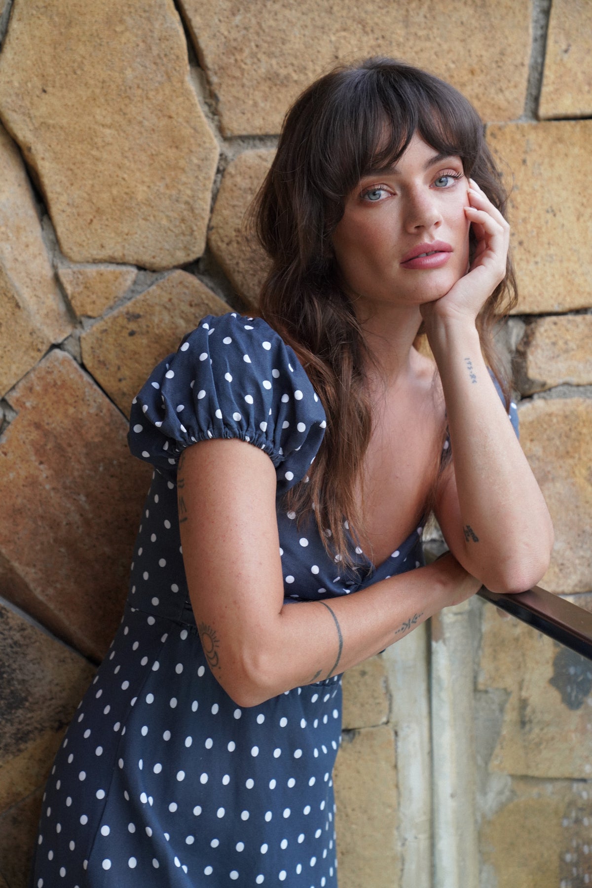 Woman in a blue polka dot dress leaning against a stone wall.