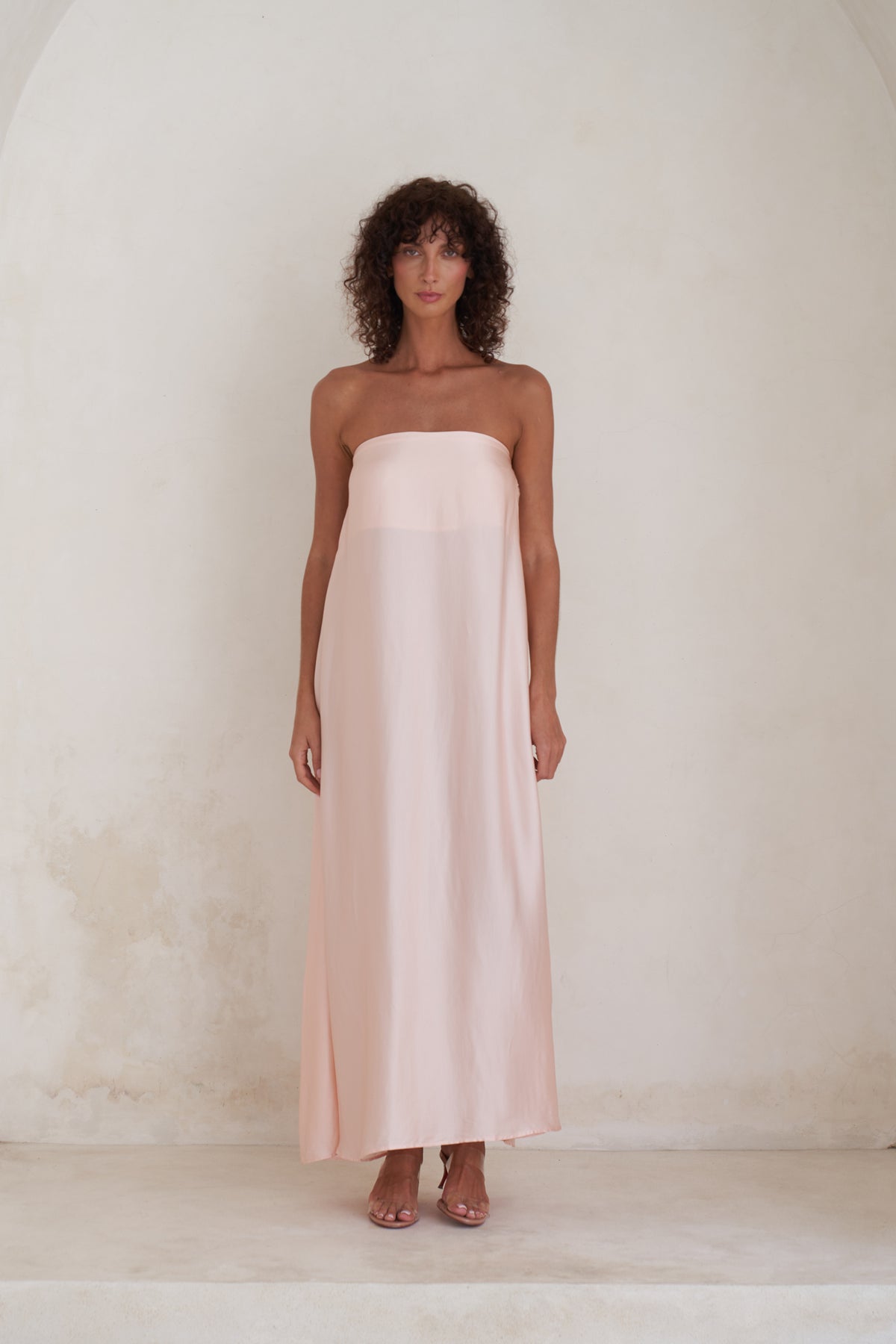 A woman stands against a textured off-white plaster wall wearing a strapless pale blush pink maxi dress with a sheer overlay.