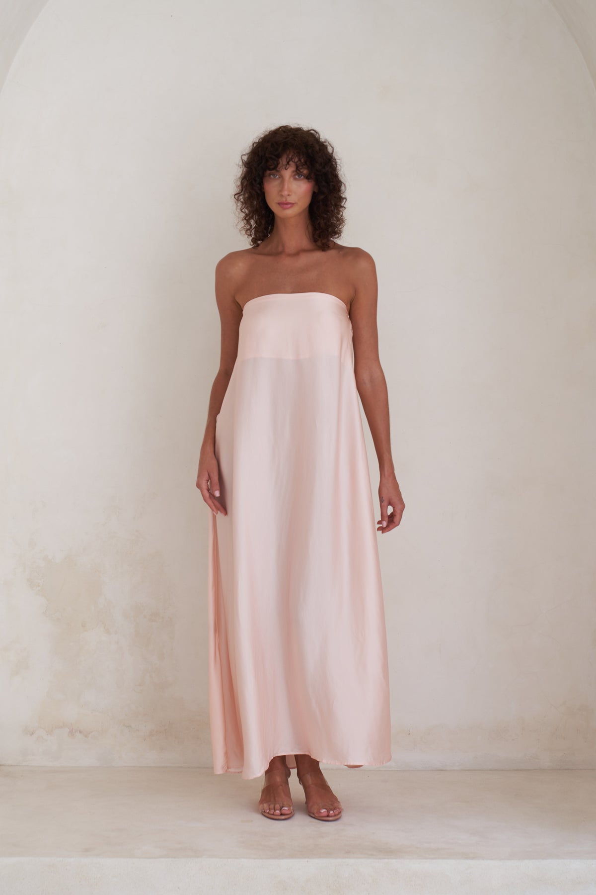 A woman wearing a strapless pale blush pink maxi dress with a draped, flowing silhouette, standing against a textured off-white plaster wall.
