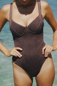 Torso of a woman with hands on her hips, she is wearing a brown one piece bathing suit