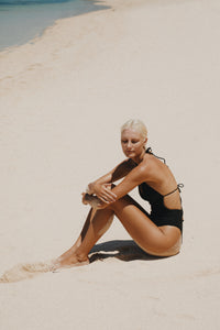 Blonde haired woman sitting on the sand wearing black halter neck one piece bathing suit