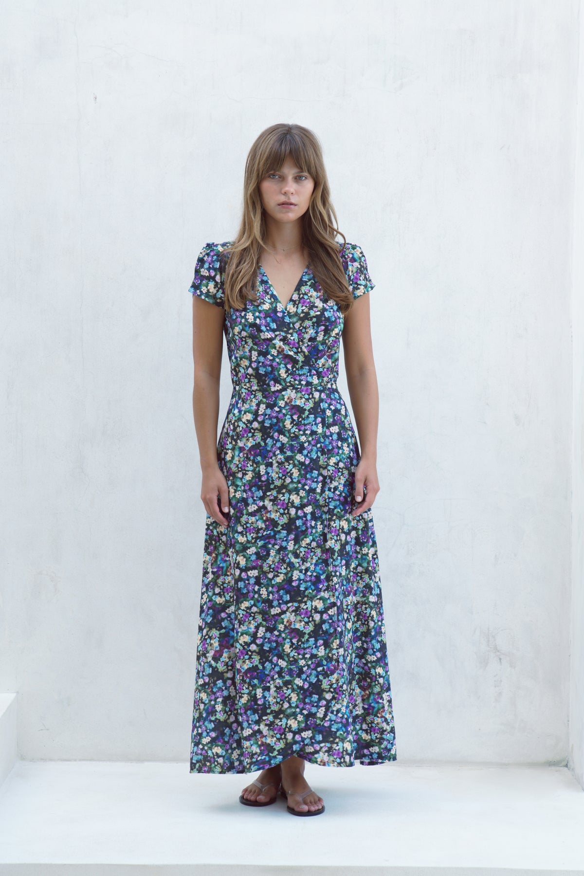 A woman stands against a white textured wall wearing a short-sleeve, V-neck maxi dress with an all-over blue, purple, and green floral print.