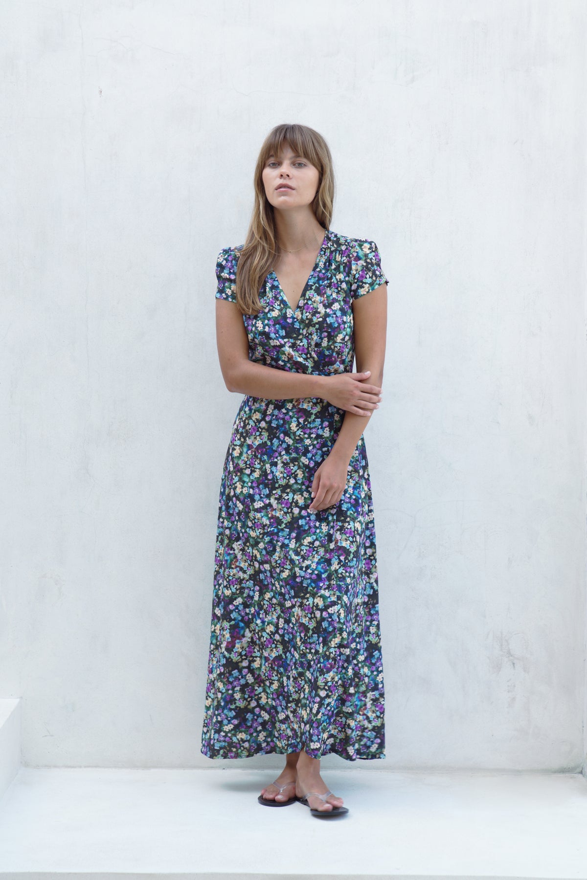 A woman stands against a white textured wall wearing a short-sleeve wrap-style maxi dress with a dense multicolor floral print in blue, green, purple, and pink tones.