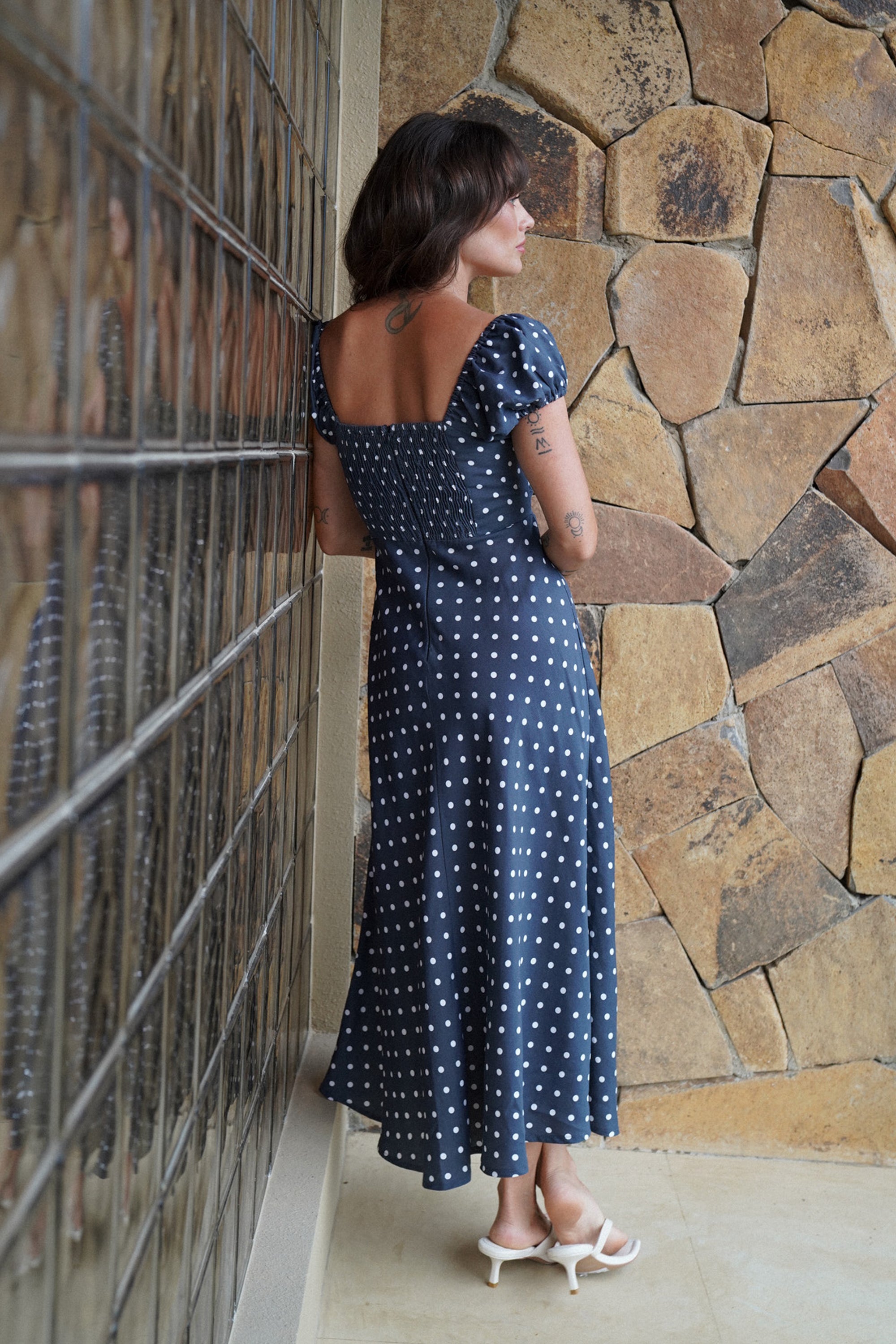 Woman in a blue polka dot dress standing against a stone wall.