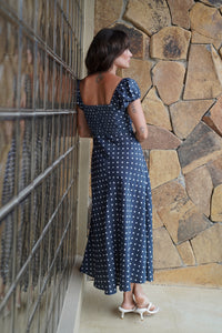 Woman in a blue polka dot dress standing against a stone wall.
