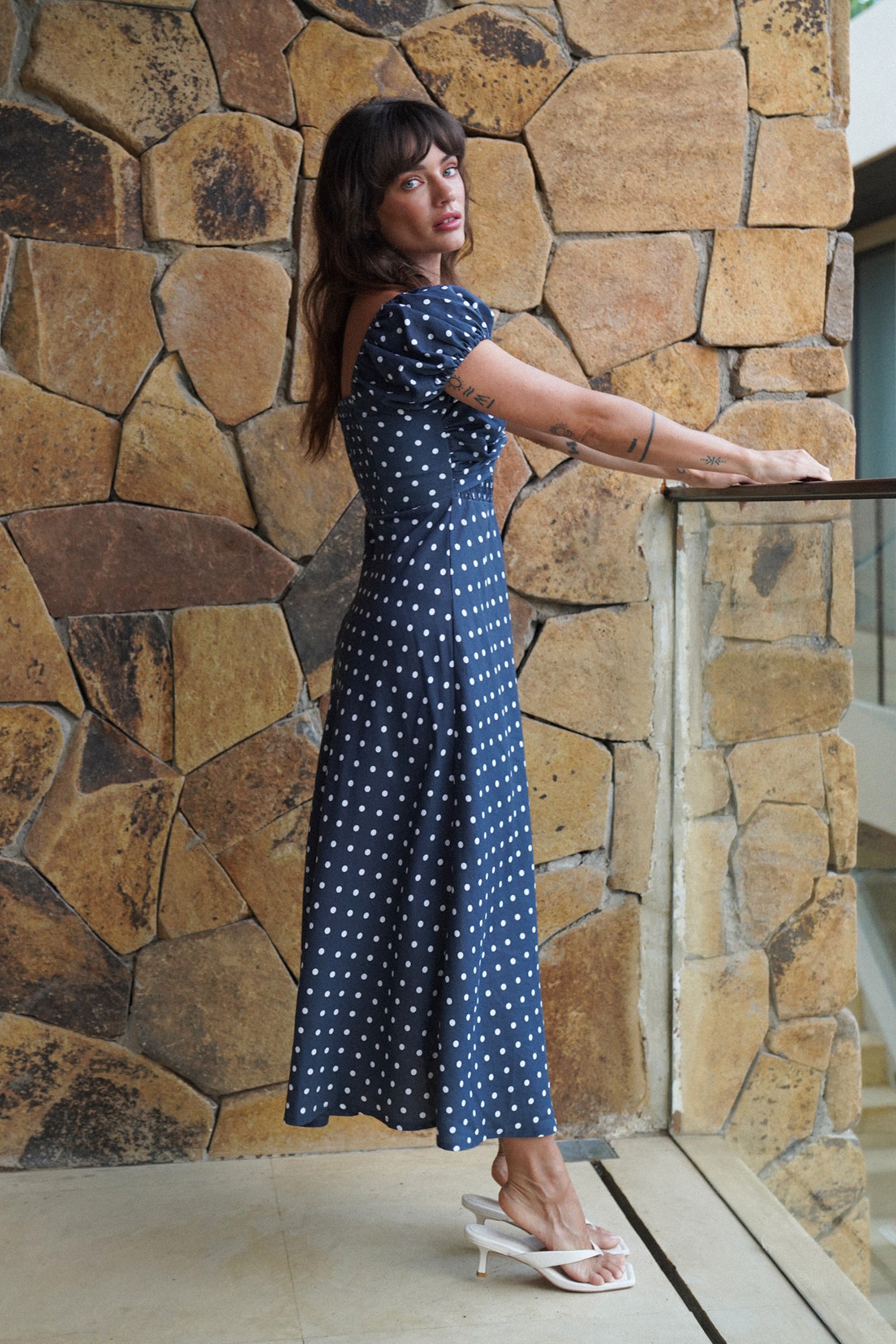 Woman in a blue polka dot dress standing against a stone wall.