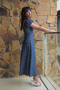 Woman in a blue polka dot dress standing against a stone wall.