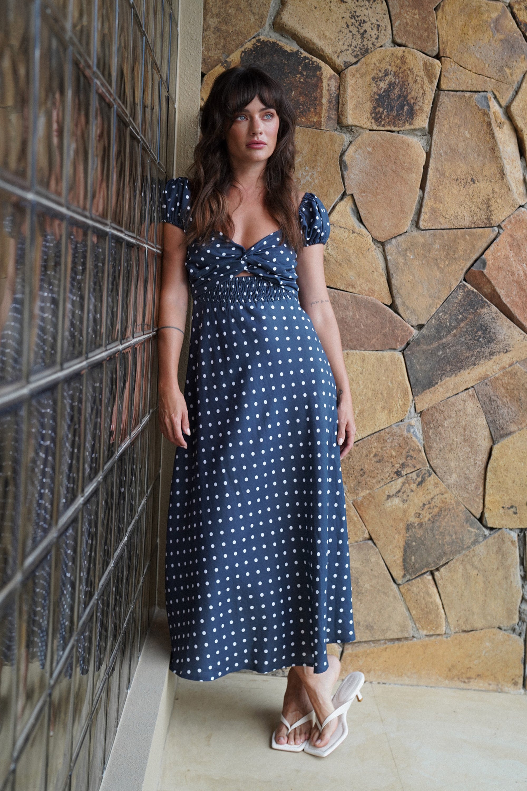 Woman in a black polka dot dress standing against a stone wall.