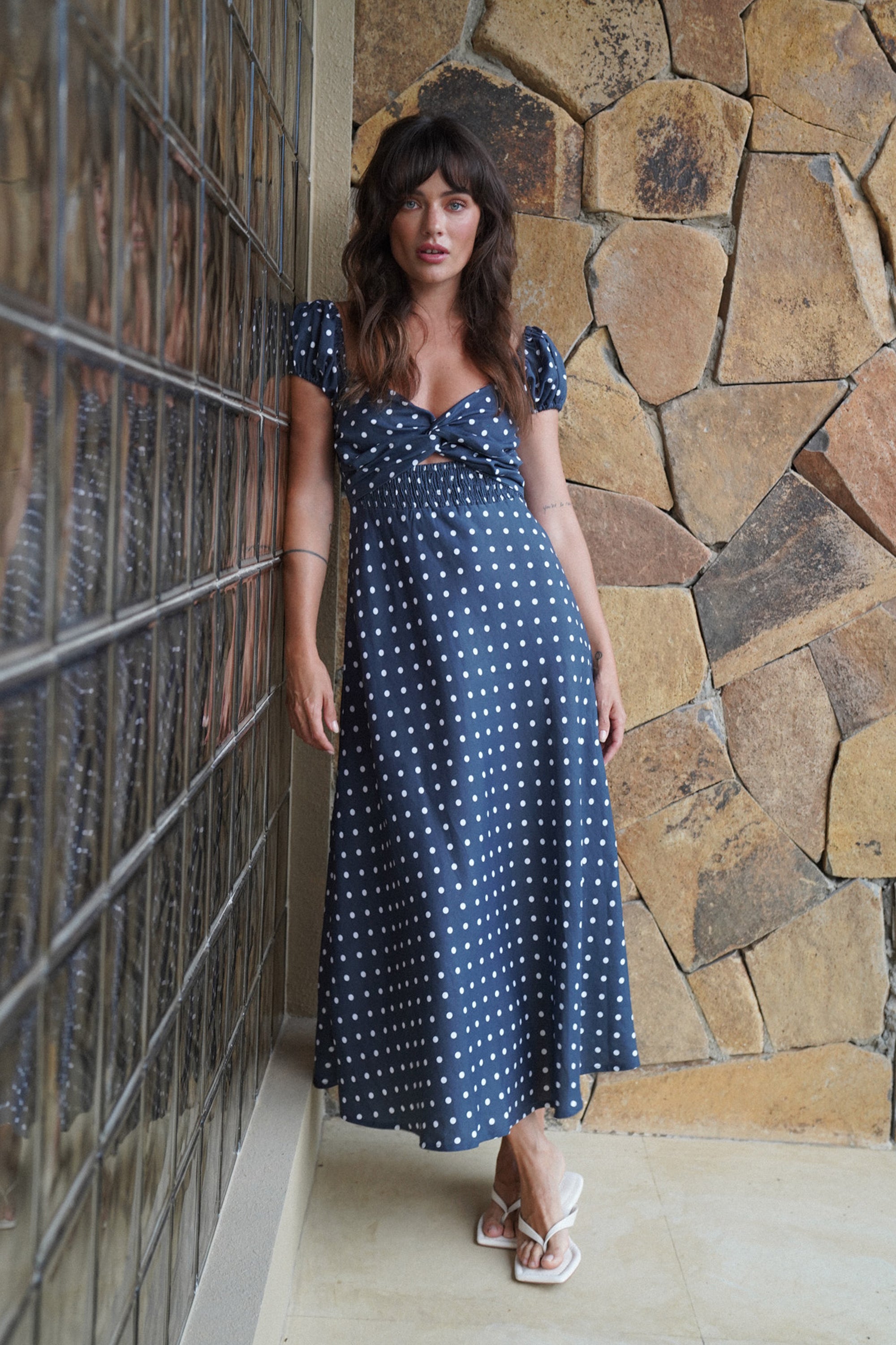 Woman in a blue polka dot dress standing against a stone wall.