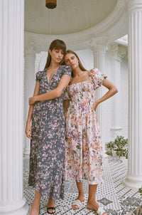 Two women standing on a porch wearing floral print maxi summer dresses  leaning on each other