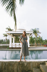 Farther view of a woman wearing square neck gingham midi dress standing in front of a pool