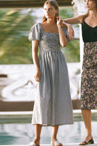 Two women looking at their side wearing gingham dress and black top with floral midi skirt standing next to a pool