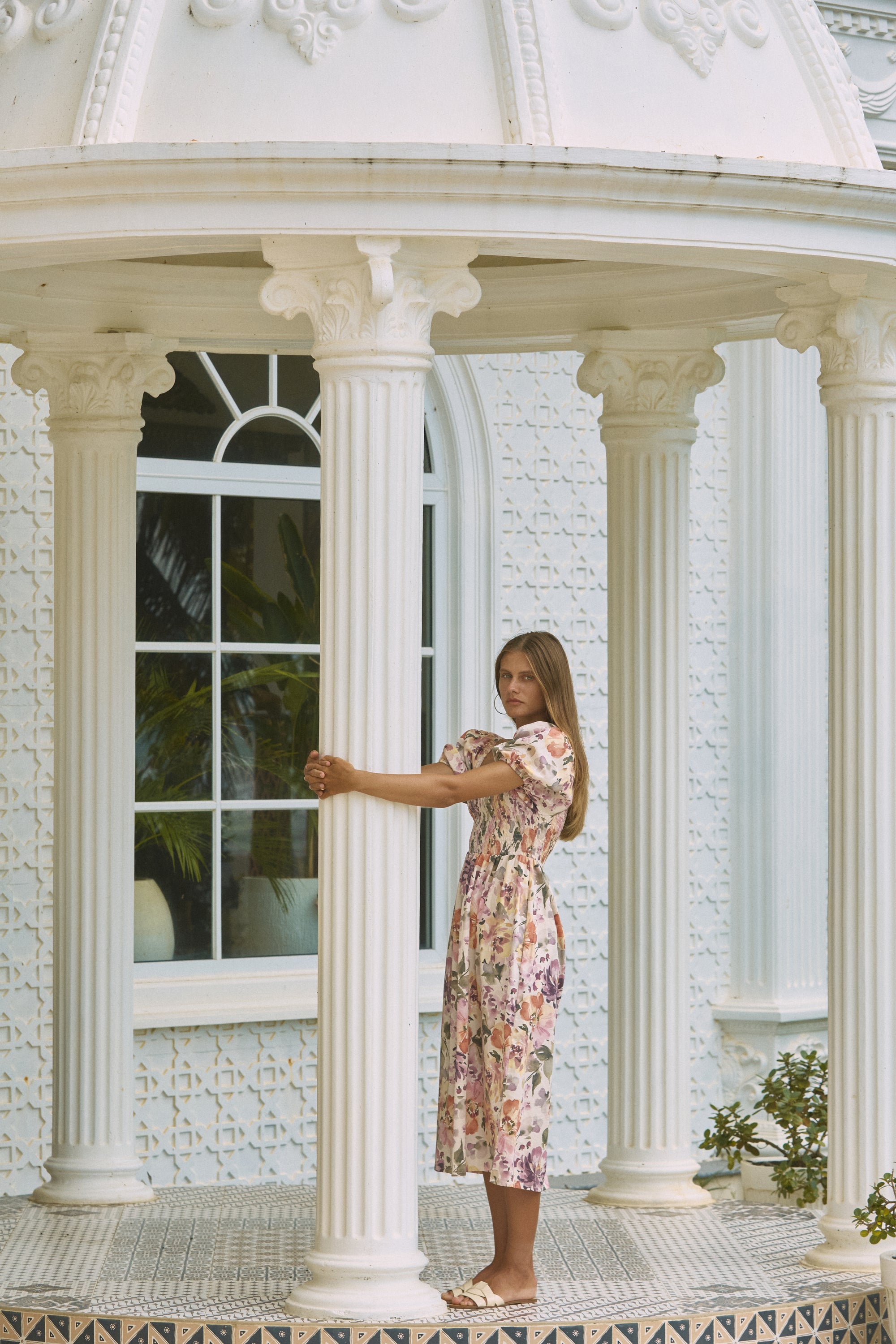 Farther view of woman wearing rosy floral printed summer dress hugging a front porch post 