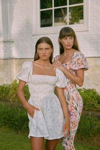 Two women modelling wearing shirred floral print dresses standing in front of a window