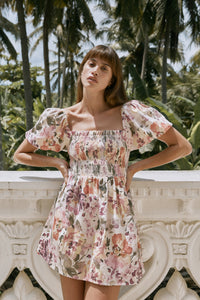 Woman with both hands in waist wearing floral printed mini dress leaning on a white balustrade