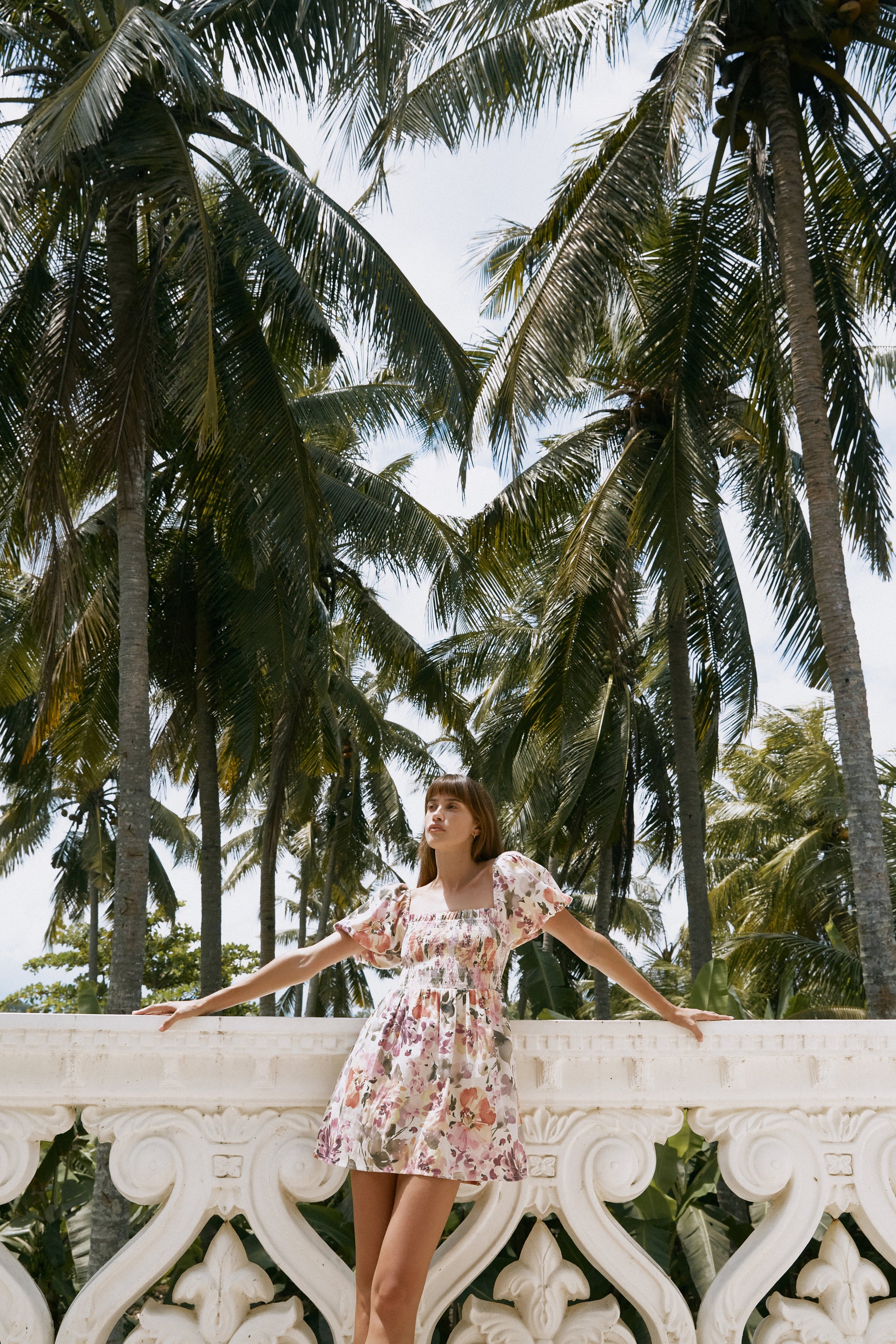 Woman wearing floral printed summer dress leaning on a white balustrade with palm tress at the background