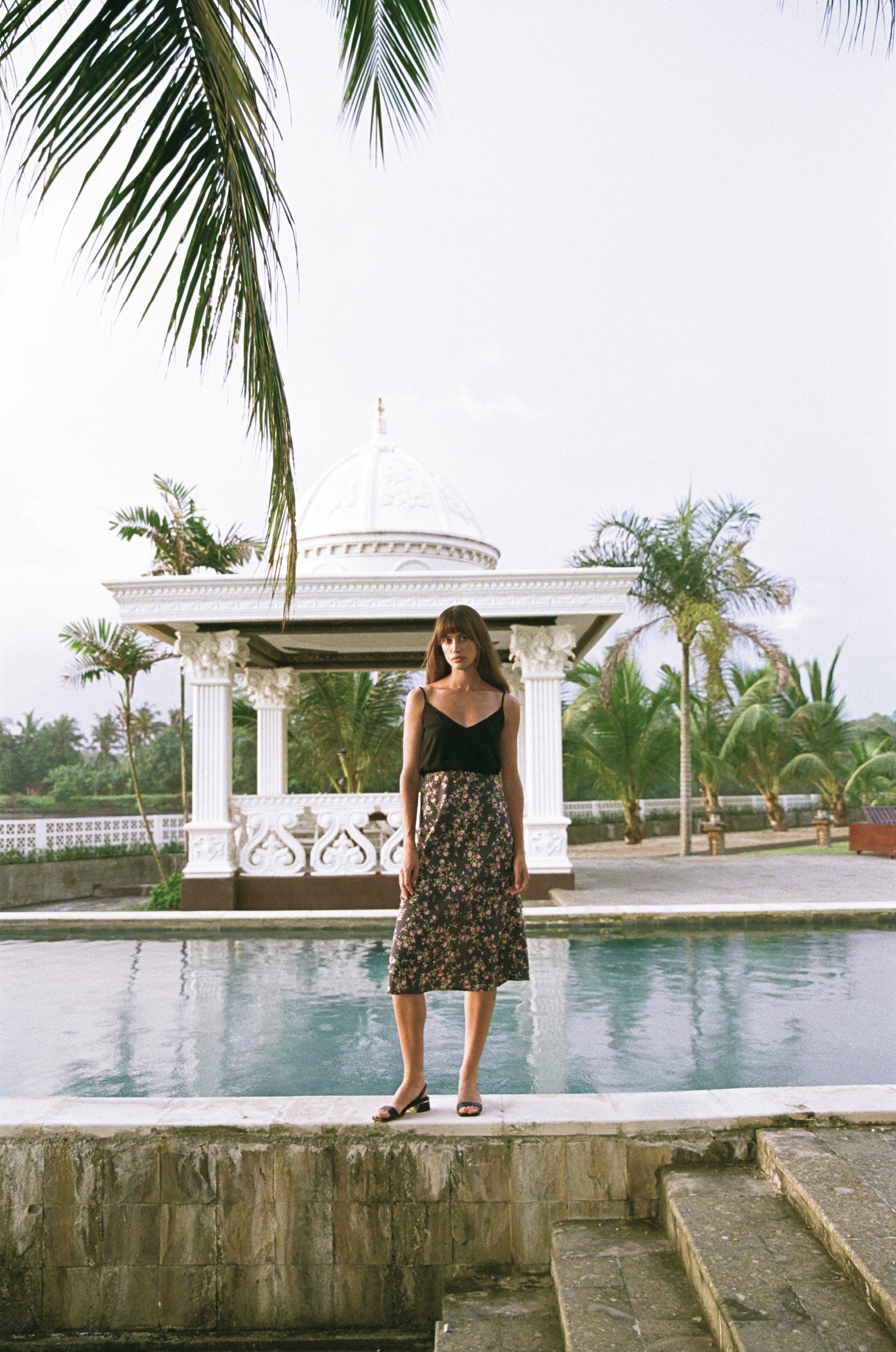 Woman wearing a black singlet top and floral print midi skirt standing next to a pool
