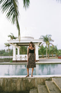Farther view of woman wearing a black singlet top and floral print midi skirt standing next to a pool