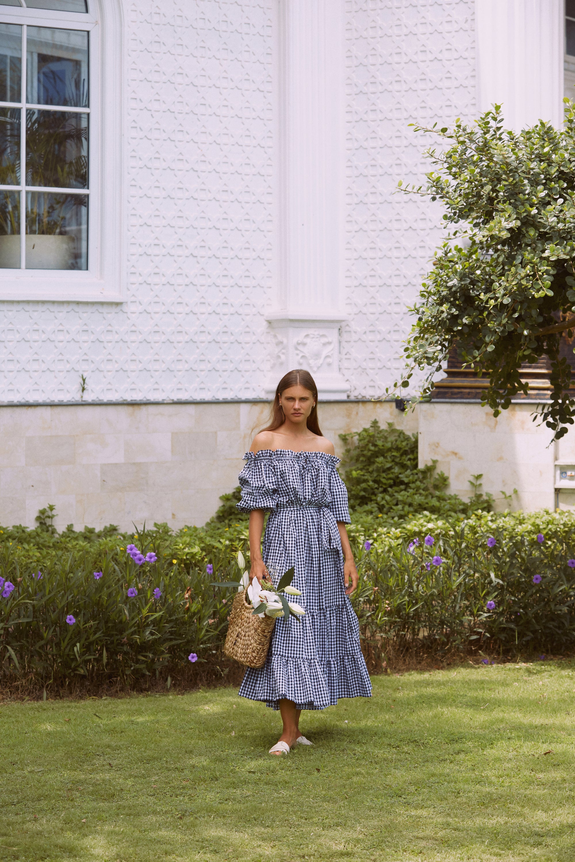 Woman in a maxi gingham dress carrying basket with flowers standing in front landscape