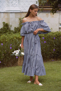 Woman in a maxi gingham dress with one hand carrying a basket with flowers and one hand in waist