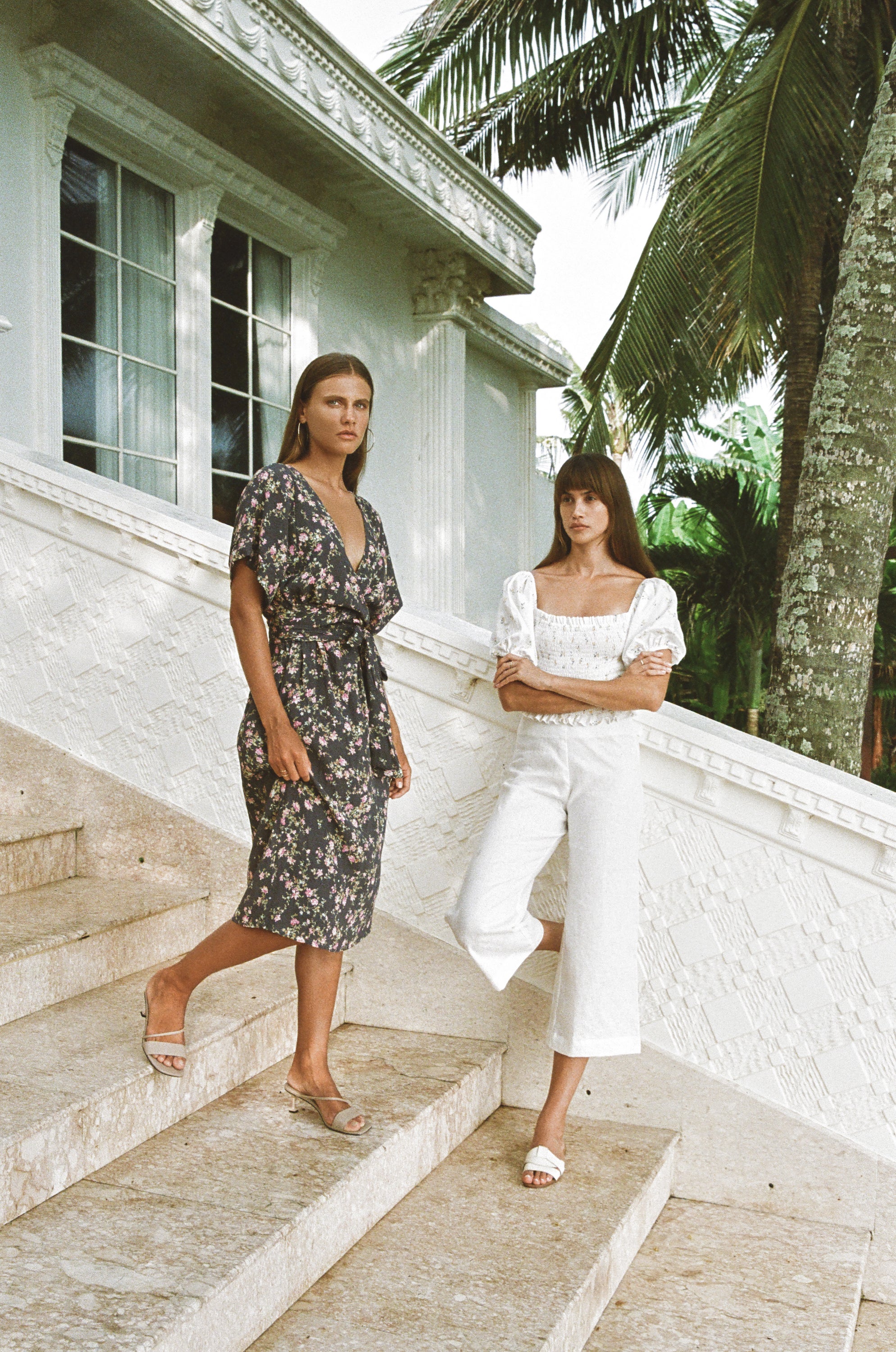 Two women wearing floral print midi dress and white floral top and pants standing on stair steps